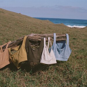 May include: Five sports bras in various colors hang on a weathered log. The bras are in shades of tan, mustard yellow, olive green, off-white, and light blue. The scene is set outdoors with a grassy hill and ocean in the background.