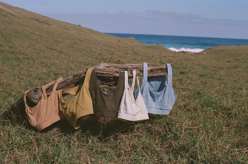 May include: Five sports bras in various colors hang on a weathered log. The bras are in shades of tan, mustard yellow, olive green, off-white, and light blue. The scene is set outdoors with a grassy hill and ocean in the background.