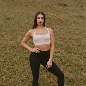 May include: A woman wearing a cream-colored sports bra and black leggings. The athletic wear is suitable for workouts. The woman is standing on a grassy field, wearing white sneakers.