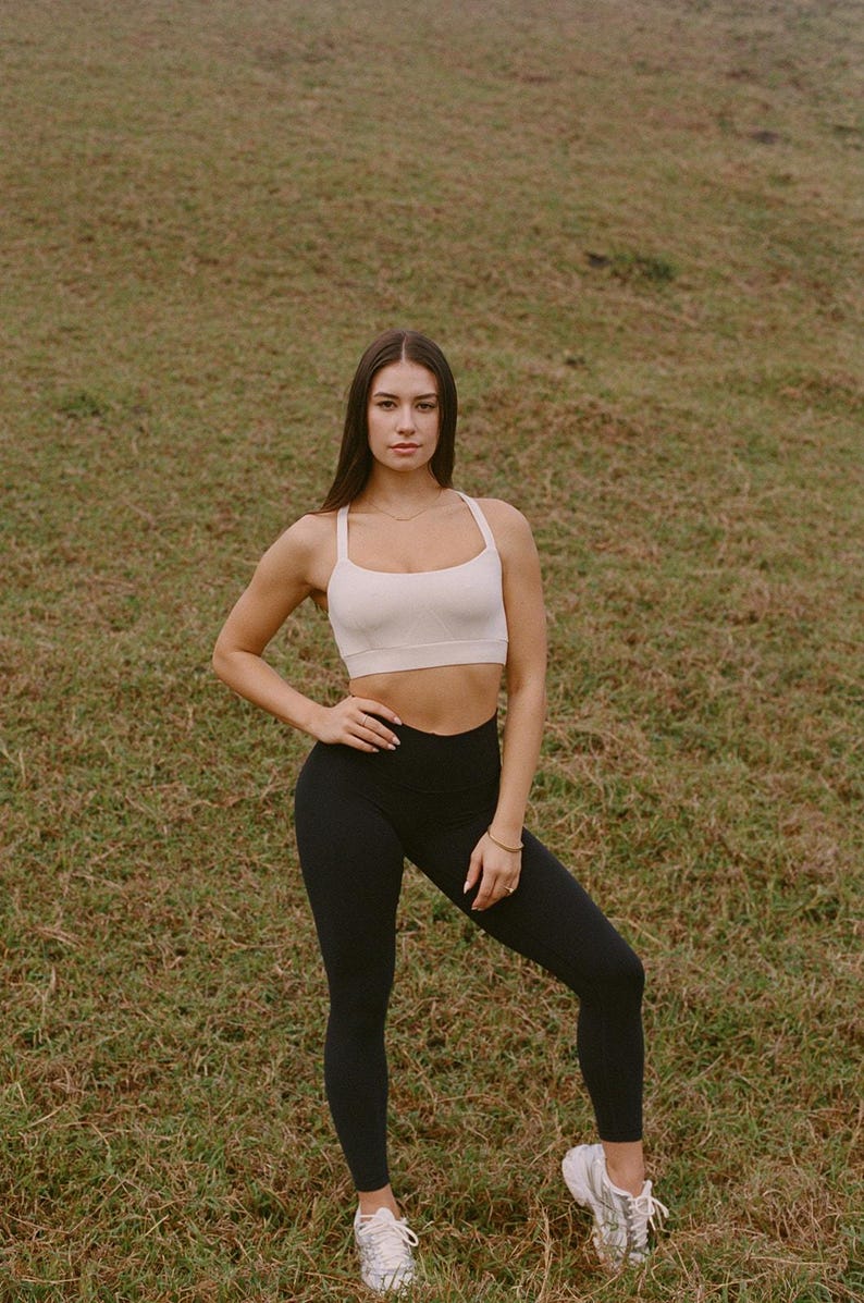 May include: A woman wearing a cream-colored sports bra and black leggings. The athletic wear is suitable for workouts. The woman is standing on a grassy field, wearing white sneakers.