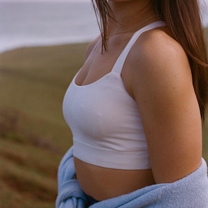 May include: A white crop top with thin straps. A light blue sweater is draped around the waist. The background features a blurred landscape with a body of water and green hills.
