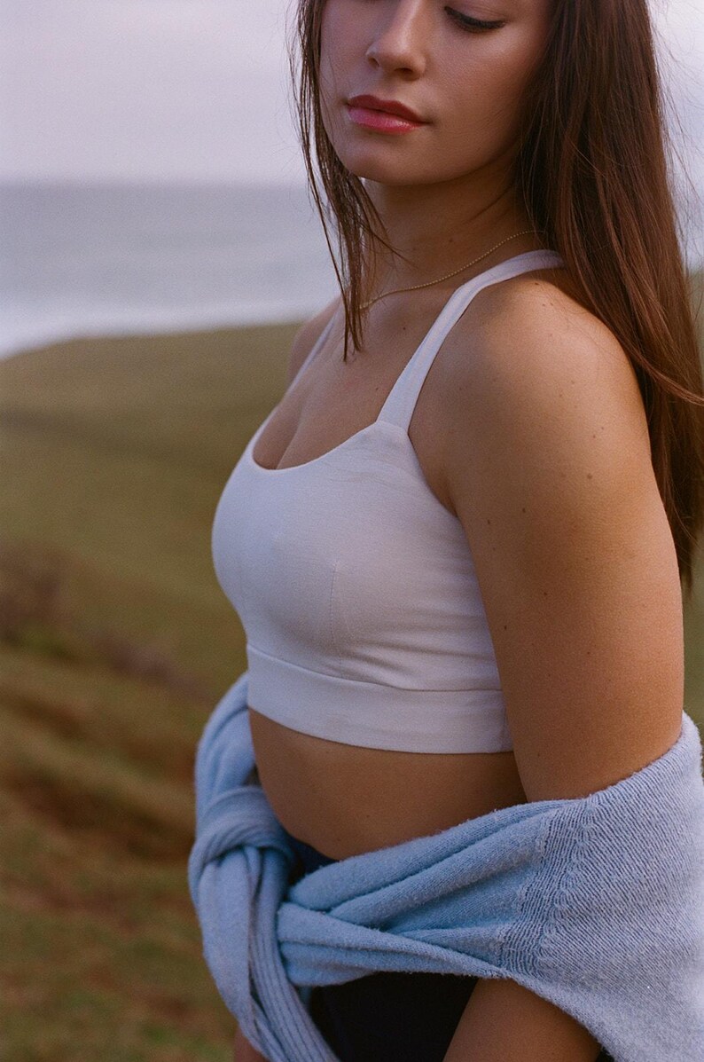May include: A white crop top with thin straps. A light blue sweater is draped around the waist. The background features a blurred landscape with a body of water and green hills.