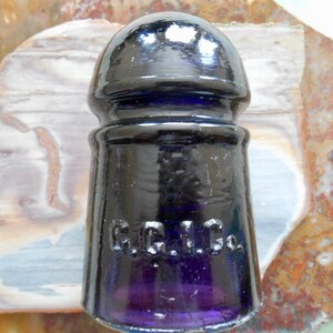 Peut inclure: Un isolateur en verre violet foncé avec un dessus arrondi et une base en forme de cloche. L'isolateur porte l'inscription en relief "G.G. Co." sur le côté. Le verre a une finition brillante et réfléchit la lumière.