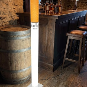 May include: A tall, cylindrical floor lamp designed to resemble a cigarette. The top section features a yellow-orange speckled filter pattern, while the lower portion is white and illuminated. The text Marlboro LIGHT is printed in black on the white section.