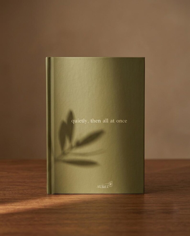 Pu&ograve; includere: Un libro rilegato con copertina verde oliva che riporta il testo "quietly, then all at once" stampato in bianco. La parte inferiore mostra il nome del marchio "AUREL" con una piccola icona a forma di fiore. L'ombra di una foglia &egrave; proiettata sulla copertina.