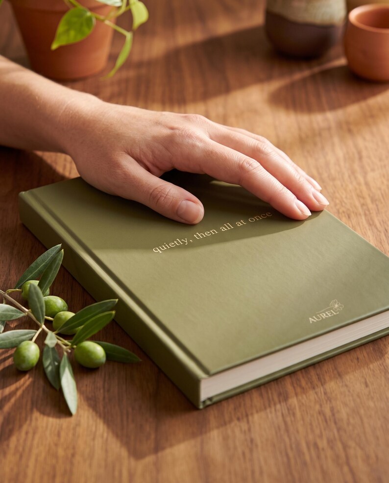 Pu&ograve; includere: Un diario con copertina rigida color verde oliva con il testo "quietly, then all at once" stampato in oro sulla copertina anteriore. Il nome del marchio "AUREL" &egrave; stampato in oro vicino all'angolo in basso a destra della copertina liscia.