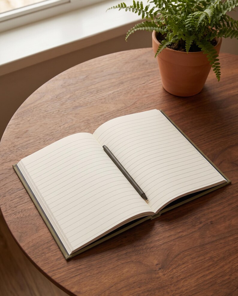 Pu&ograve; includere: Un taccuino aperto con pagine color crema a righe &egrave; appoggiato in piano, caratterizzato da una copertina verde oliva scuro. Una penna abbinata verde oliva scuro &egrave; posizionata in diagonale al centro delle pagine aperte, pronta per scrivere.