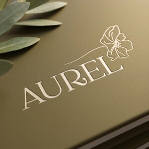 Pu&ograve; includere: Una copertina di libro verde oliva opaco con la parola AUREL impressa in un colore crema chiaro. Un delicato disegno al tratto di un singolo fiore &egrave; posizionato sopra la lettera L, e un piccolo rametto di foglie d'olivo verdi poggia nell'angolo in alto a sinistra.