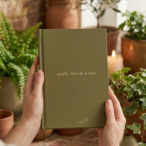 Pu&ograve; includere: Un diario rettangolare con copertina rigida color verde oliva con il testo 'quietly, then all at once' stampato in oro sulla copertina anteriore. Il nome del marchio 'AUREL' &egrave; stampato in oro vicino al centro della parte inferiore.