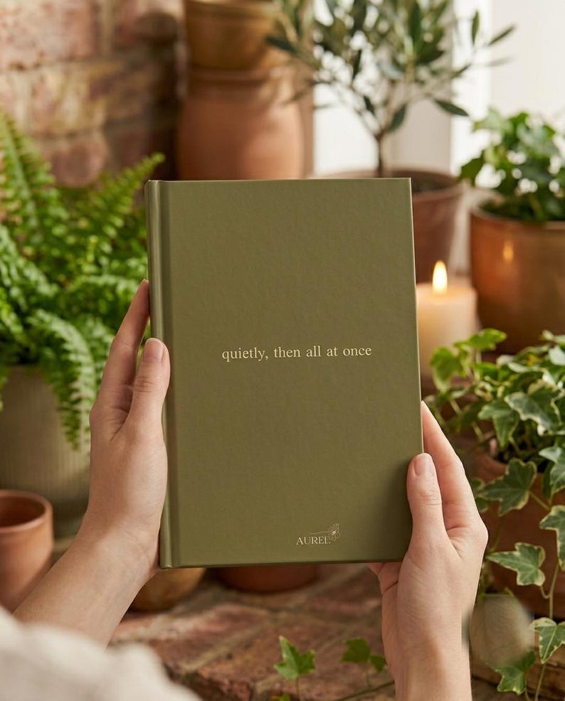 Pu&ograve; includere: Un diario rettangolare con copertina rigida color verde oliva con il testo 'quietly, then all at once' stampato in oro sulla copertina anteriore. Il nome del marchio 'AUREL' &egrave; stampato in oro vicino al centro della parte inferiore.