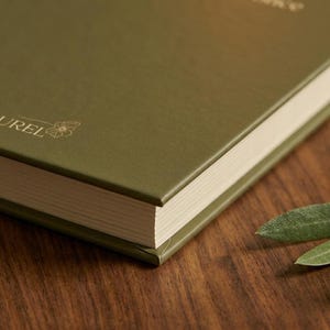 Pu&ograve; includere: Un libro con copertina rigida verde oliva con una finitura opaca liscia. La copertina presenta un testo in lamina d'oro che recita "then all at once" e la parola "LAUREL" accompagnata da un piccolo disegno al tratto di un fiore. Pagine color crema.