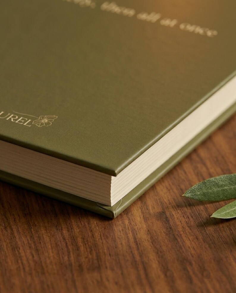 Pu&ograve; includere: Un libro con copertina rigida verde oliva con una finitura opaca liscia. La copertina presenta un testo in lamina d'oro che recita "then all at once" e la parola "LAUREL" accompagnata da un piccolo disegno al tratto di un fiore. Pagine color crema.