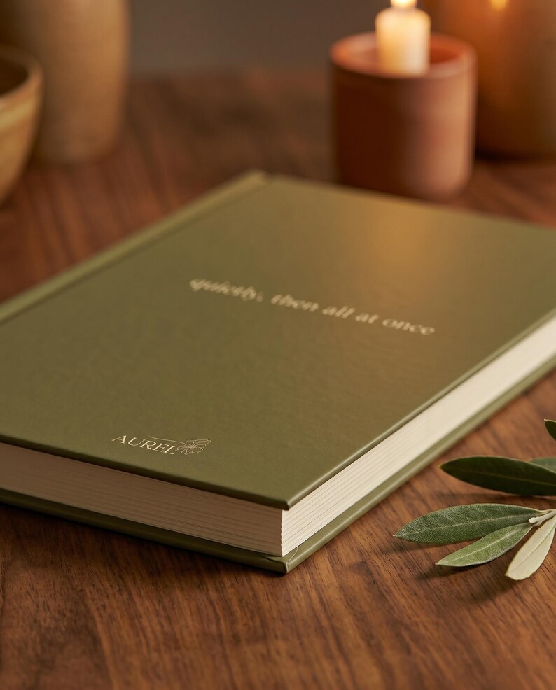 Pu&ograve; includere: Un libro rilegato con copertina verde oliva che presenta il testo "quietly, then all at once" impresso in oro al centro e il nome del marchio "AUREL" con una piccola icona floreale stampata in oro in basso a sinistra sulla copertina.