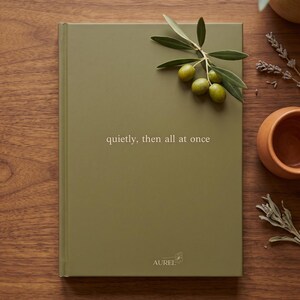 Pu&ograve; includere: Un taccuino con copertina rigida verde oliva opaco con il testo 'quietly, then all at once' stampato in bianco. Al centro in basso &egrave; riportato il nome del marchio 'AUREL' con una piccola icona floreale. Un rametto di olive verdi &egrave; appoggiato sopra.