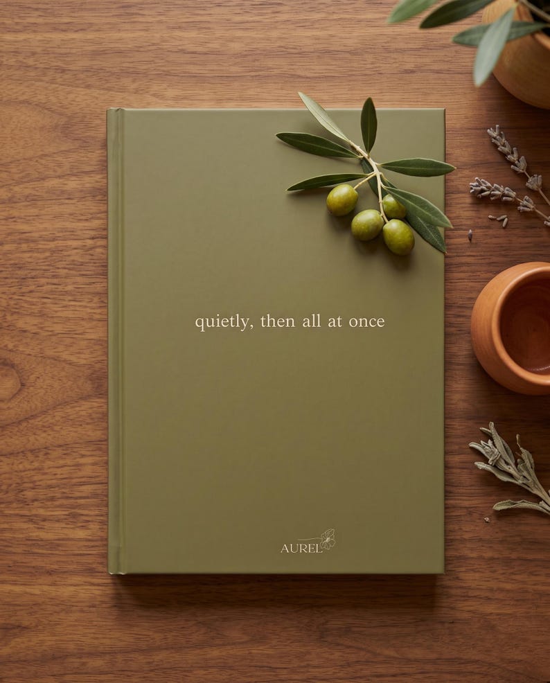 Pu&ograve; includere: Un taccuino con copertina rigida verde oliva opaco con il testo 'quietly, then all at once' stampato in bianco. Al centro in basso &egrave; riportato il nome del marchio 'AUREL' con una piccola icona floreale. Un rametto di olive verdi &egrave; appoggiato sopra.