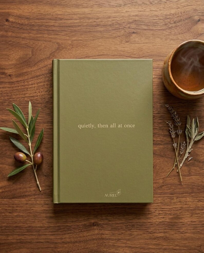 Pu&ograve; includere: Un quaderno con copertina rigida verde oliva con il testo "quietly, then all at once" stampato in oro sulla copertina anteriore. La parte inferiore della copertina riporta il marchio "AUREL" in piccole lettere dorate con un disegno di un fiore.
