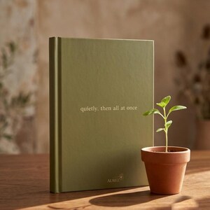 Pu&ograve; includere: Un libro con copertina rigida color verde oliva con il testo "quietly, then all at once" stampato in bianco sulla copertina anteriore, insieme alla parola "AUREL" vicino al bordo inferiore. Una piccola pianta verde in un vaso di terracotta.