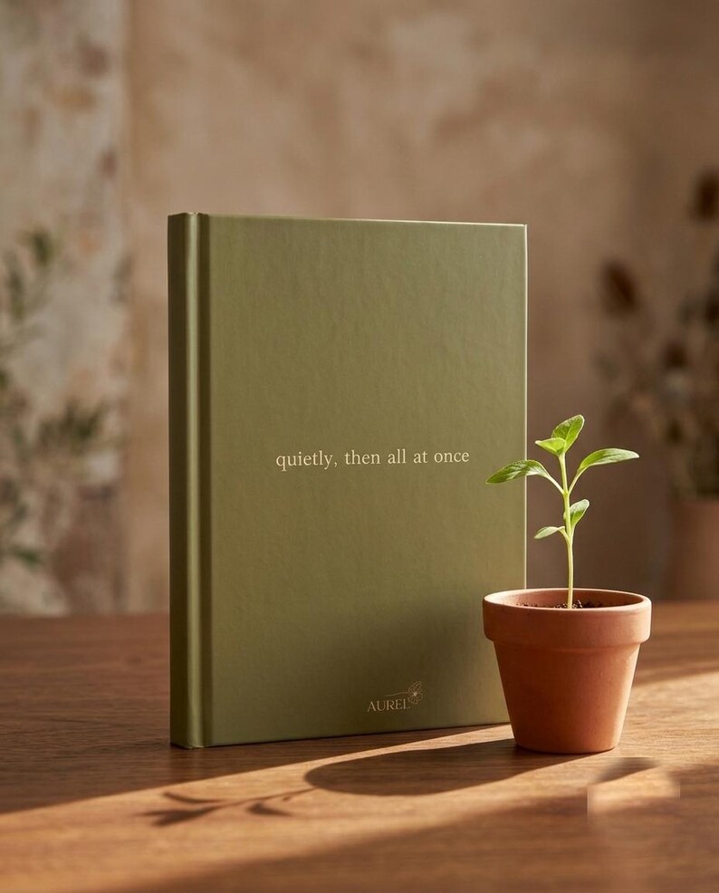 Pu&ograve; includere: Un libro con copertina rigida color verde oliva con il testo "quietly, then all at once" stampato in bianco sulla copertina anteriore, insieme alla parola "AUREL" vicino al bordo inferiore. Una piccola pianta verde in un vaso di terracotta.