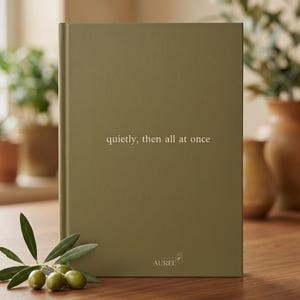 Pu&ograve; includere: Un libro rettangolare con copertina rigida color verde oliva con il testo "quietly, then all at once" stampato in oro sulla copertina. La parola "AUREL" e una piccola icona di un fiore sono stampate in oro vicino al centro inferiore.