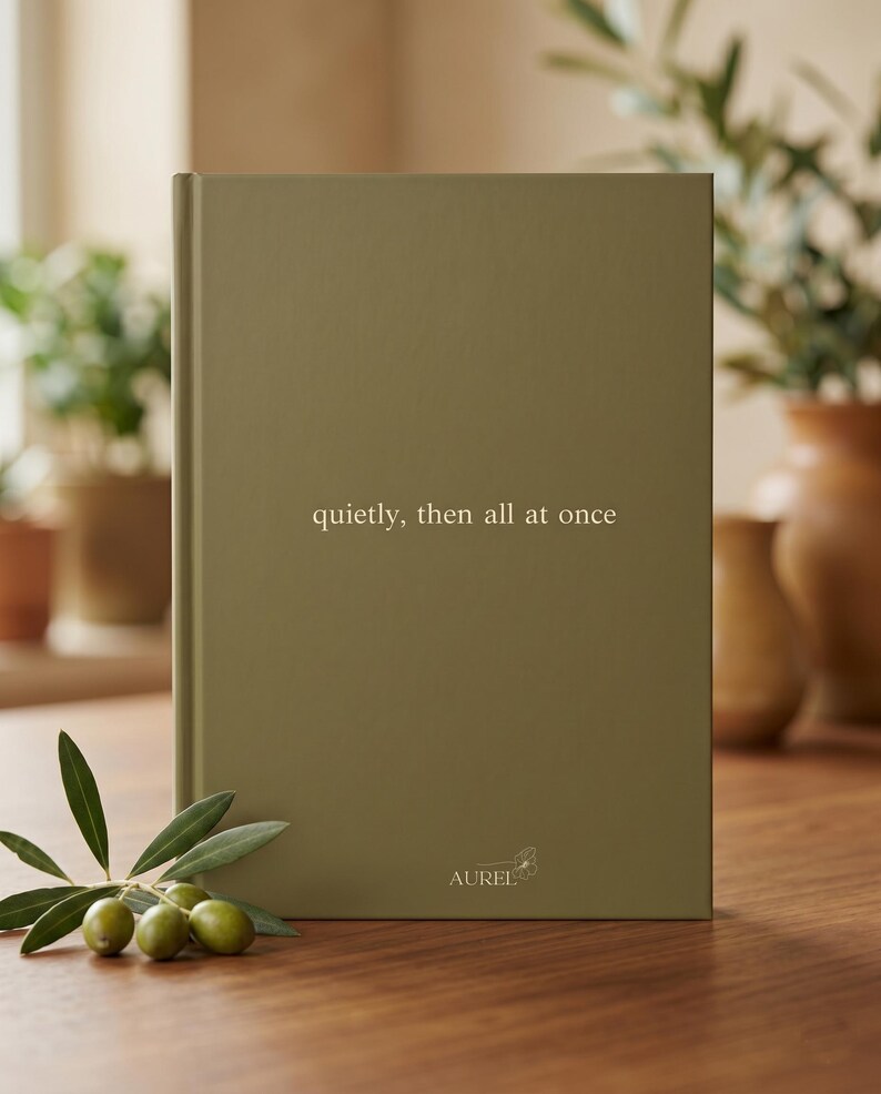 Pu&ograve; includere: Un libro rettangolare con copertina rigida color verde oliva con il testo "quietly, then all at once" stampato in oro sulla copertina. La parola "AUREL" e una piccola icona di un fiore sono stampate in oro vicino al centro inferiore.