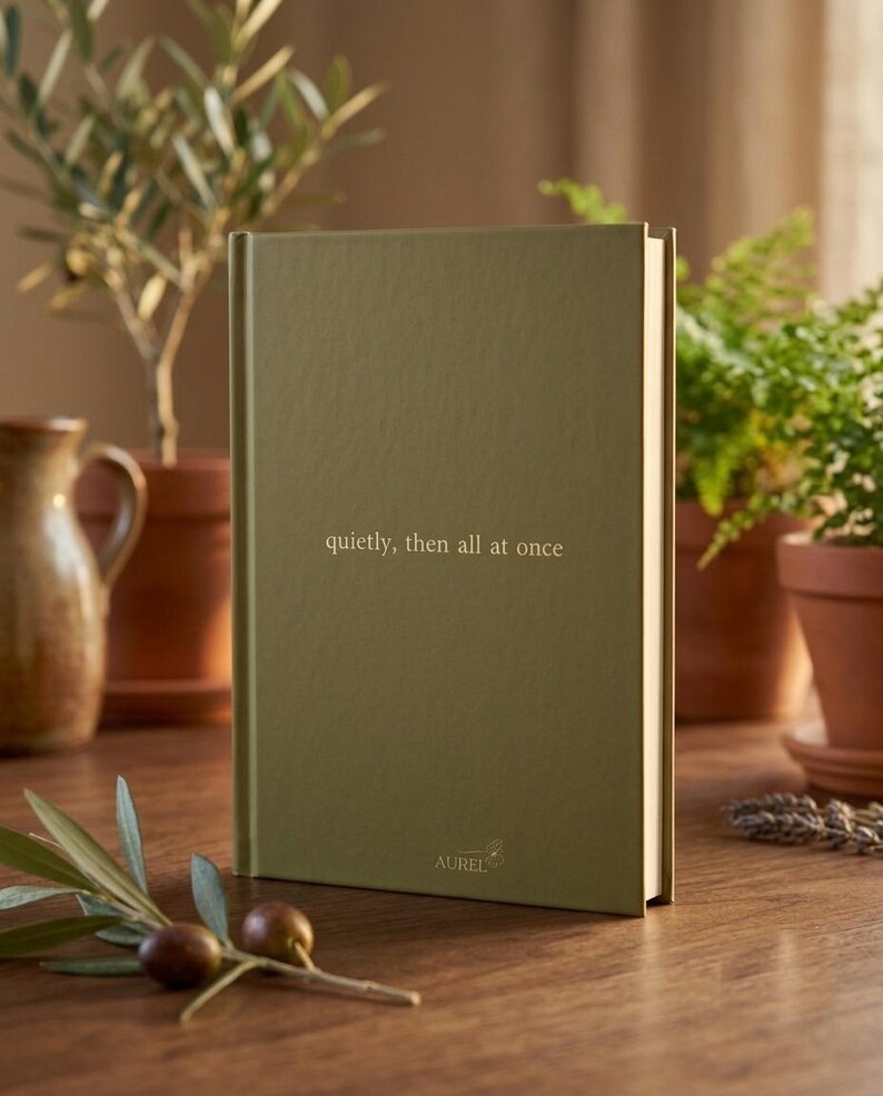 Pu&ograve; includere: Un libro con copertina rigida di colore verde oliva opaco, caratterizzato dal testo 'quietly, then all at once' impresso in oro al centro e dal nome del marchio 'AUREL' stampato in oro vicino al bordo inferiore della copertina.