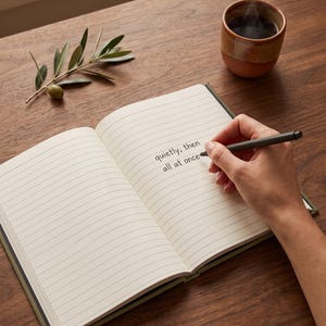 Pu&ograve; includere: Un taccuino aperto con pagine a righe che presenta il testo scritto a mano 'quietly, then all at once' in inchiostro nero. Una penna grigio scuro &egrave; tenuta sopra la pagina. Nelle vicinanze si trovano una piccola tazza in ceramica e un ramo d'ulivo.