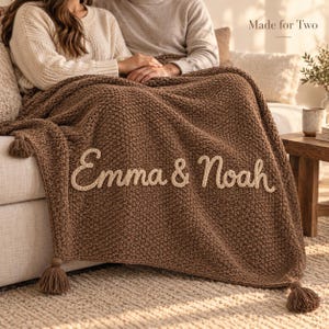 Puede incluir: Una manta de punto marrón con el texto bordado en color crema "Emma & Noah" en el centro. La manta tiene un patrón de punto grueso y texturizado, y está rematada con borlas marrones a juego en las esquinas para un toque decorativo.