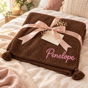 Op de afbeelding: Een donkerbruine gebreide deken met een textuurpatroon en kwastjes aan de hoeken. De deken is gestrikt met een lichtroze satijnen lint, bevat een klein leeg wit cadeaulabel en heeft de naam Penelope in roze borduursel op de voorzijde.