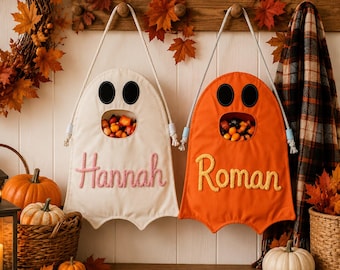 May include: Two fabric ghost-shaped wall hangings with rope handles, one white with the name Hannah in pink and one orange with the name Roman in yellow. Each ghost features black oval eyes and a circular mouth opening filled with colorful candy.