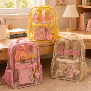 Könnte beinhalten: Drei durchsichtige Rucksäcke mit seitlichen Netztaschen und aufgestickten Namen auf der Vorderseite. Der gelbe Rucksack trägt den Namen Emilee, der rosa Rucksack Olivia und der beige Rucksack den Namen Grace, alle mit Schreibwaren gefüllt.