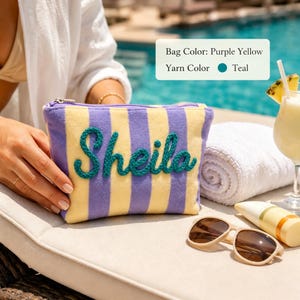 Op de afbeelding: Een rechthoekig tasje met ritssluiting en verticale paarse en gele strepen. De naam Sheila is op de voorzijde geborduurd met groenblauw garen. Een tekstvak rechtsboven toont de tekst: Bag Color: Purple Yellow, Yarn Color: Teal.