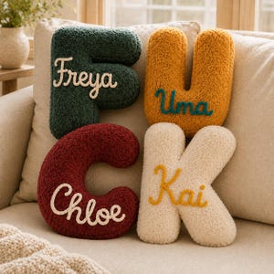 May include: Four plush letter-shaped cushions in dark green, mustard yellow, deep red, and cream. The green F features the name Freya, the yellow U features Uma, the red C features Chloe, and the cream K features the name Kai in embroidered script.