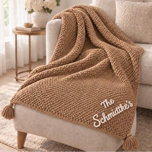 May include: A tan knitted throw blanket with a textured waffle pattern and tassels on the corners. The blanket features white embroidered text that reads The Schmidtke's, positioned in the lower right corner of the fabric.