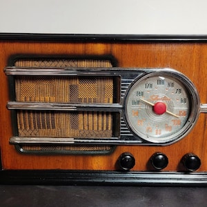 May include: A rectangular tabletop radio with a wood grain finish and black trim. It features a brown fabric speaker grille, a circular silver dial with a red center knob, and three black control knobs along the bottom. The dial displays the text Silvertone.