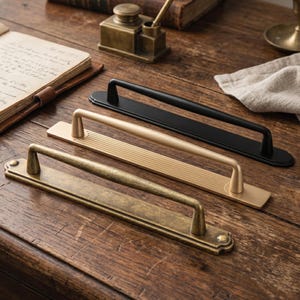 May include: Three elongated cabinet pulls arranged in a row, featuring a matte black finish, a brushed gold finish with a ribbed texture on the base plate, and an antique brass finish with a weathered, textured surface on the base plate.