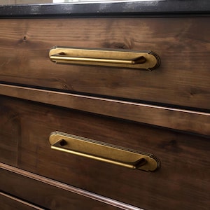 May include: Two horizontal brass drawer pulls with a rectangular backplate featuring rounded corners and a smooth finish. The hardware is mounted onto dark brown wooden drawers with visible grain patterns, creating a contrast between the metal and wood.