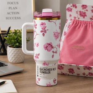 May include: White and pink floral print tumbler with a handle. The tumbler has a pink lid and gold trim. The text "QUENCHER H2.0 TUMBLER" is printed on the side. A pink drawstring bag and floral box are in the background.