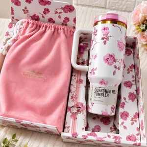 May include: A white tumbler with pink rose floral pattern, a handle, and a pink lid, presented in a decorative box with matching floral design. Includes a pink drawstring bag and straw. The tumbler has the text "QUENCHER H2.0 TUMBLER".