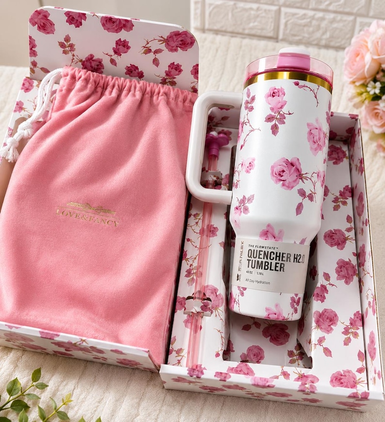 May include: A white tumbler with pink rose floral pattern, a handle, and a pink lid, presented in a decorative box with matching floral design. Includes a pink drawstring bag and straw. The tumbler has the text "QUENCHER H2.0 TUMBLER".