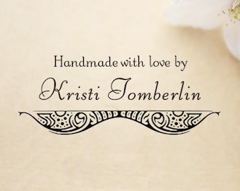 May include: A black ink stamp design on a light beige background featuring the text Handmade with love by Kristi Tomberlin in a serif font, positioned above a decorative, symmetrical horizontal flourish with intricate swirling patterns.