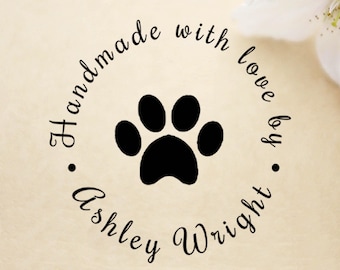 May include: A circular design with a black paw print in the center, surrounded by the words "Handmade with love by Ashley Wright" in a script font. The background is a light beige color.