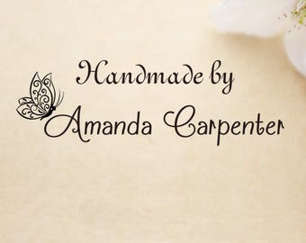 May include: A black ink stamp design featuring a decorative butterfly with intricate filigree wings positioned to the left of the text Handmade by Amanda Carpenter written in a flowing script font on a light beige textured paper background.
