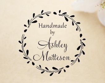 May include: A black ink rubber stamp impression featuring a circular wreath of leaves surrounding the text Handmade by Ashley Matteson. The design is printed in a clear, elegant font with a decorative script style for the name, centered on a light background.