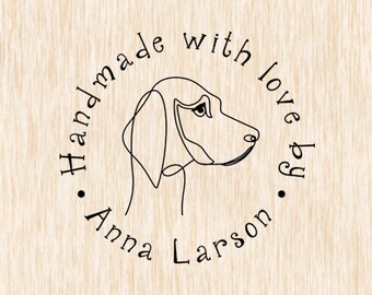 May include: A circular logo with a line drawing of a dog's head in black. The words "Handmade with love by Anna Larson" are in a circular font around the dog image. The background is a light wood grain.