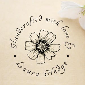 Könnte beinhalten: Ein kreisförmiges Blumendesign mit dem Text "Handcrafted with love by Laura Hedge". Das Bild zeigt eine detaillierte Schwarz-Weiß-Blumenillustration auf beigem Hintergrund, mit einer weißen Blume in der oberen rechten Ecke.