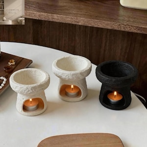 May include: Three ceramic oil burners with a rough, textured finish. Two burners are off-white and one is black. Each burner features a wide top bowl and a hollow base containing a small, lit tea light candle, creating a warm, glowing effect.