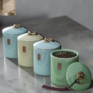 Pode incluir: Quatro latas de chá cilíndricas de cerâmica com acabamento em esmalte craquelado em tons de azul claro, amarelo pálido e verde sálvia. Cada lata possui uma tampa correspondente com alça de anel em tom de latão, uma borla castanha e um rótulo.