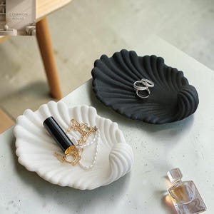 May include: Two shell-shaped trinket dishes, one matte black and one matte white, with ribbed textures. The black dish holds two silver rings. The white dish contains a gold-toned chain necklace and a black lipstick tube. A glass perfume bottle sits nearby.