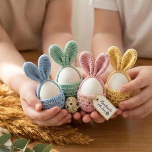 May include: Four pastel-colored crochet egg holders shaped like bunnies, each holding a white egg. The colors are blue, green, pink, and yellow. One holder has a tag that says "Handmade with Love".