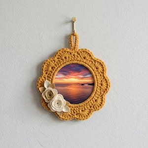 May include: A round, mustard-yellow crocheted frame with a sunset landscape image. The frame has a scalloped edge and is adorned with two fabric flowers in cream and tan. It hangs from a gold hook on a white wall.